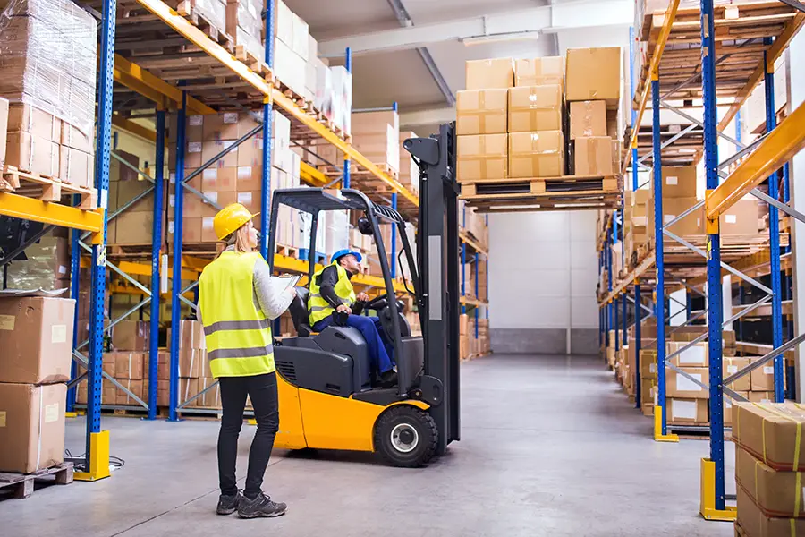 A warehouse worker operates a forklift, while another looks on