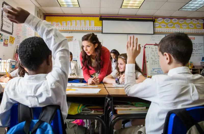 A teacher leans over her desk, holding a discussion with pupils